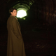 Jessie Buckley wears a long coat and stands in a tunnel, looking over her shoulder, in a scene from Men.